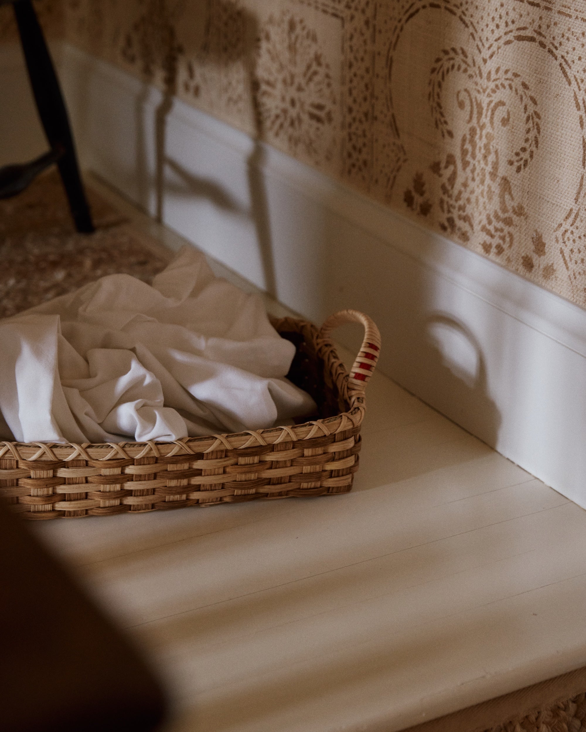 The Folk Fleur Breakfast Tray, handwoven and adorned with a white cloth, sits on a white wooden floor near patterned wallpaper.