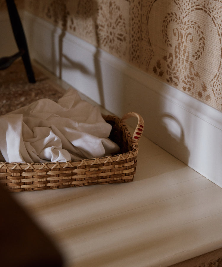 The Folk Fleur Breakfast Tray, handwoven and adorned with a white cloth, sits on a white wooden floor near patterned wallpaper.