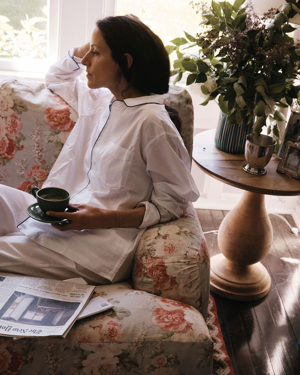 A woman in white pajamas sits on a floral couch, holding a cup and saucer. On the round wooden table beside her are a newspaper, flowers in a vase, a framed photo, and the handmade Natalia Water Jug.