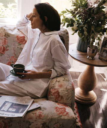 A woman in white pajamas sits on a floral couch, holding a cup and saucer. On the round wooden table beside her are a newspaper, flowers in a vase, a framed photo, and the handmade Natalia Water Jug.
