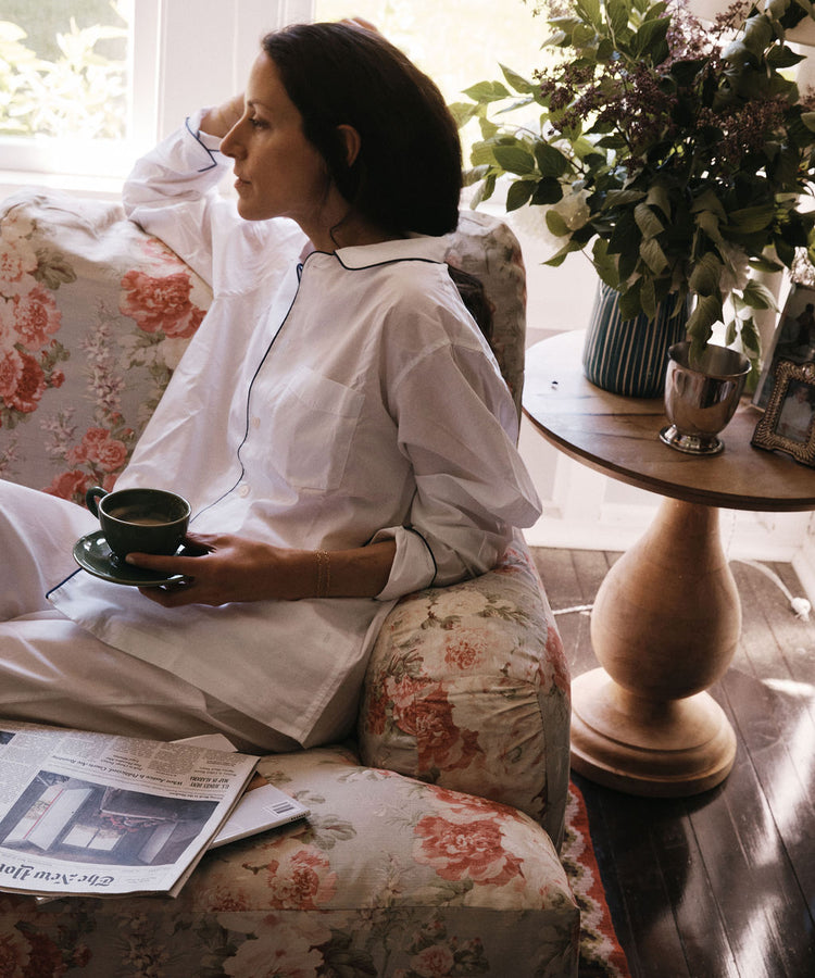 A woman in white pajamas sits on a floral couch, holding a cup and saucer. On the round wooden table beside her are a newspaper, flowers in a vase, a framed photo, and the handmade Natalia Water Jug.