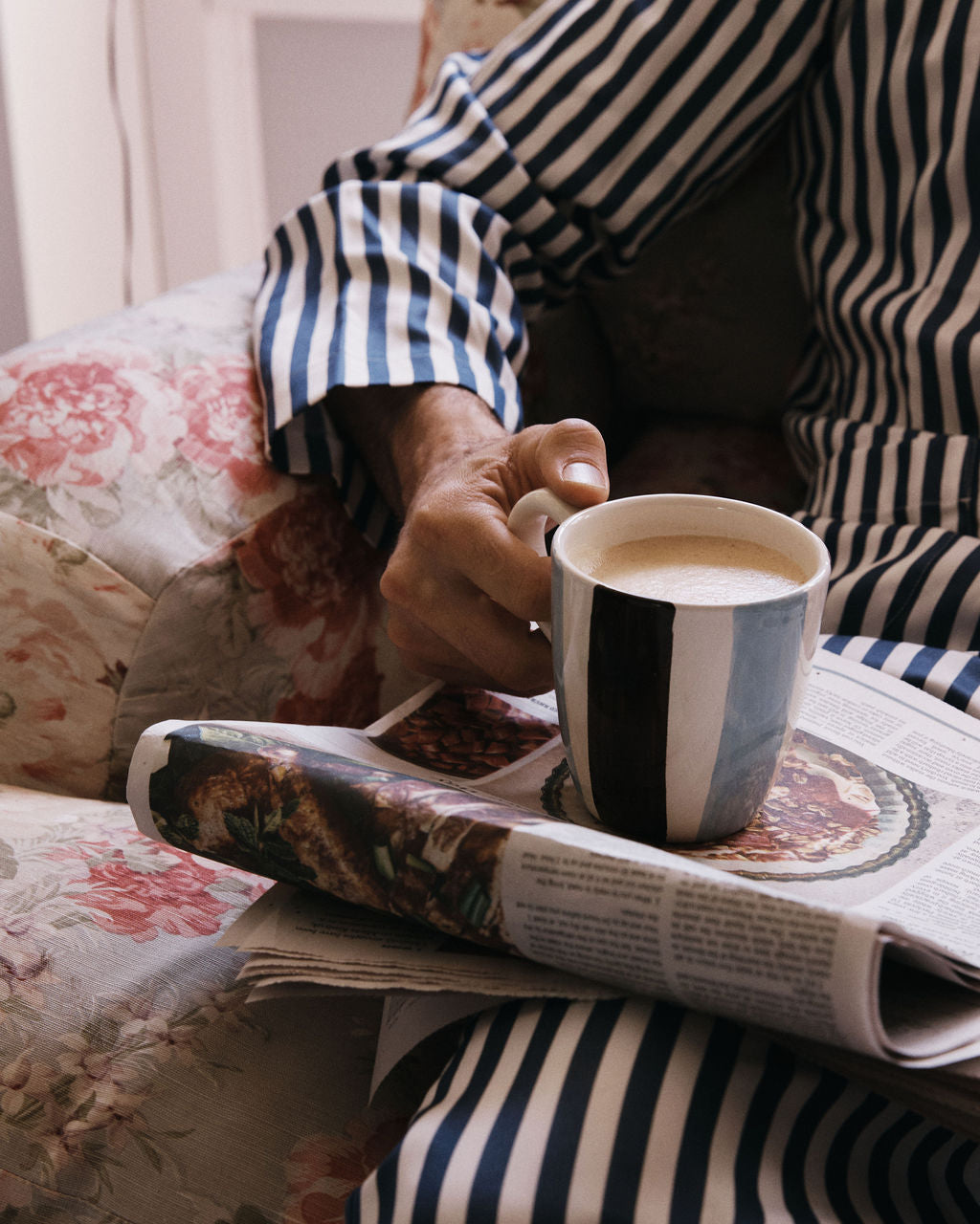 A person in striped pajamas sits on a floral armchair, holding a Mina Mug of coffee with a folded newspaper resting on their lap.