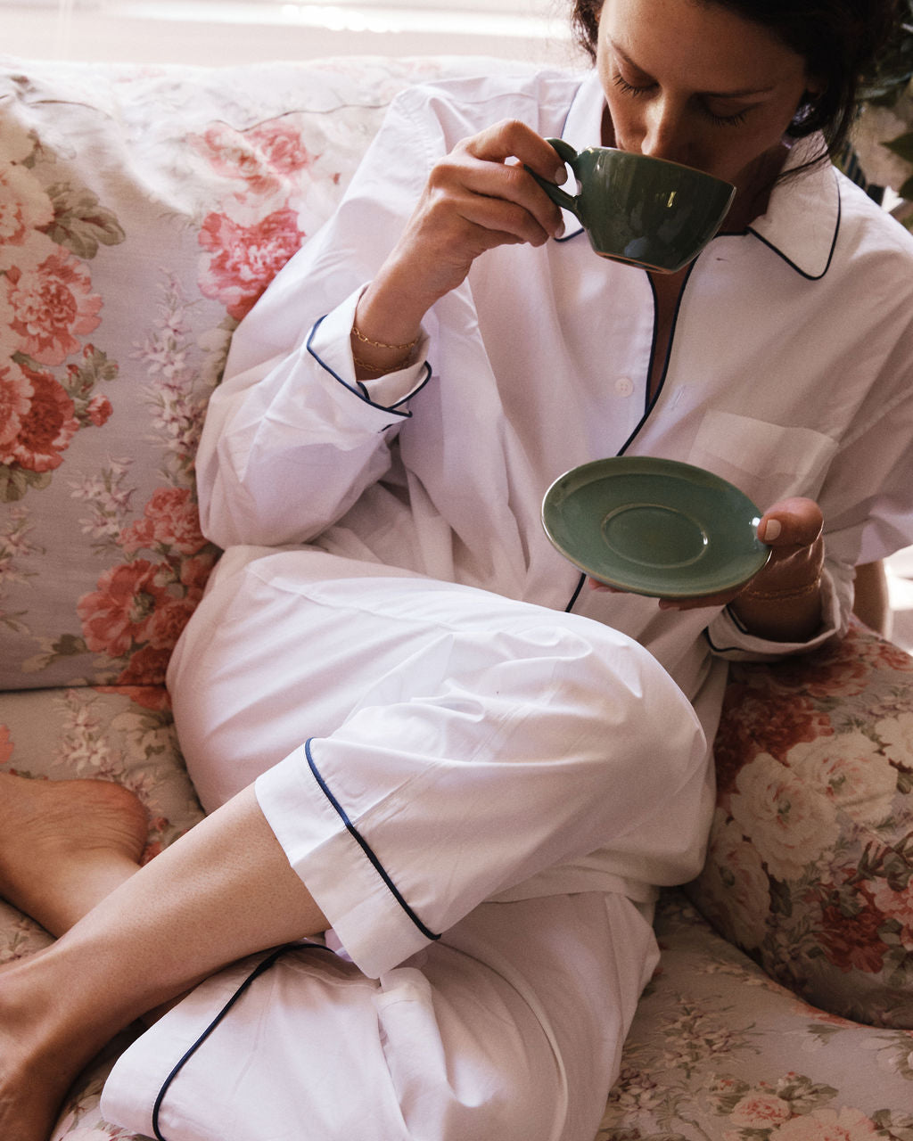 Wearing white pajamas with dark piping, a person relaxes on a floral couch, sipping from the Guido Coffee Set's cup and saucer, evoking the feel of a tranquil morning at an Italian café.