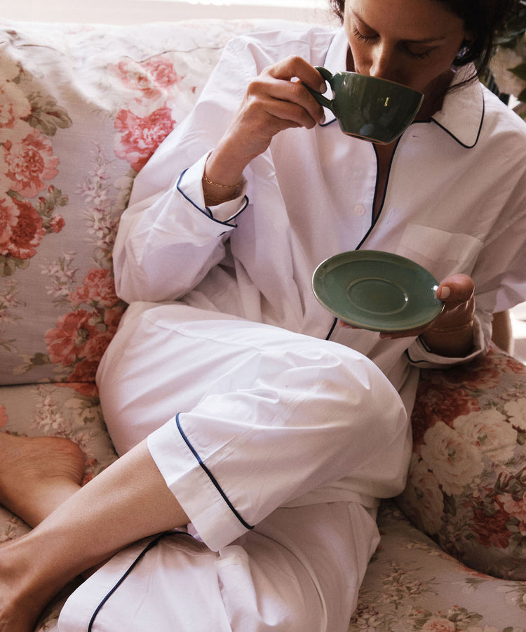 Wearing white pajamas with dark piping, a person relaxes on a floral couch, sipping from the Guido Coffee Set's cup and saucer, evoking the feel of a tranquil morning at an Italian café.
