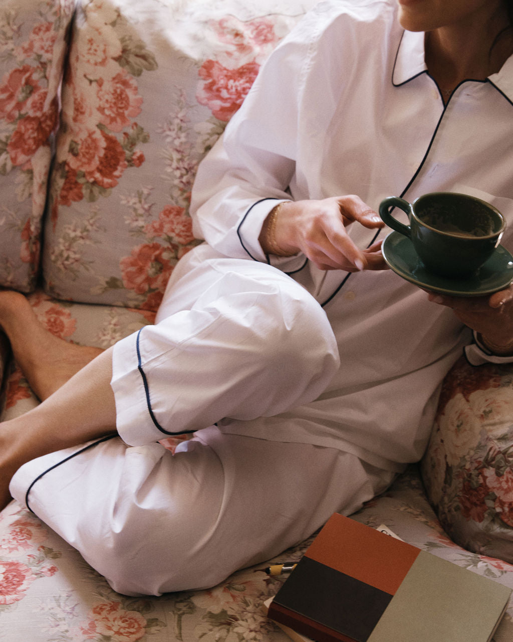 A person in white pajamas, holding a cup and saucer, sits on a floral-patterned couch beside two closed books, ready to organize thoughts in the nearby Dreamer's Notebook with pen to paper.