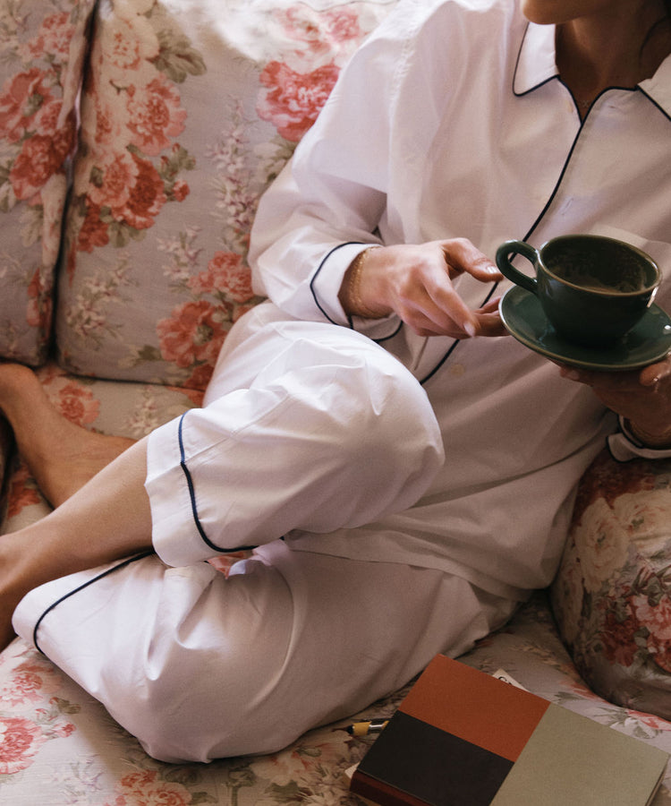 A person in white pajamas, holding a cup and saucer, sits on a floral-patterned couch beside two closed books, ready to organize thoughts in the nearby Dreamer's Notebook with pen to paper.
