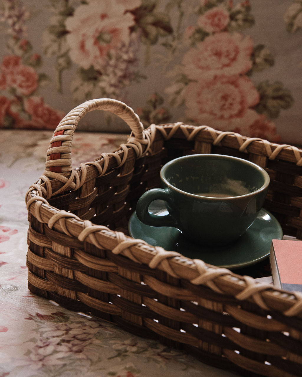 The Folk Fleur Breakfast Tray features a green cup and saucer in a woven reed basket, with a closed book and floral-patterned fabric adding charm to the scene.
