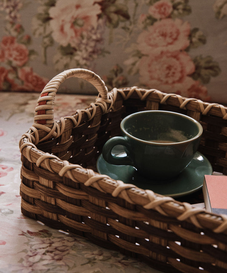 The Folk Fleur Breakfast Tray features a green cup and saucer in a woven reed basket, with a closed book and floral-patterned fabric adding charm to the scene.