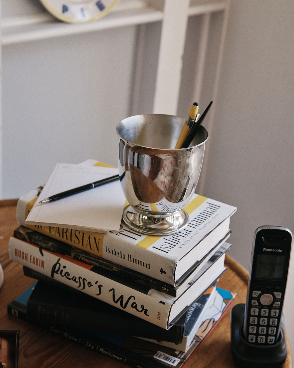 A stack of books, a notepad, a pen, a Lena Pen Pot holding pencils like a pewter vase, and a cordless phone rest on a wooden table.