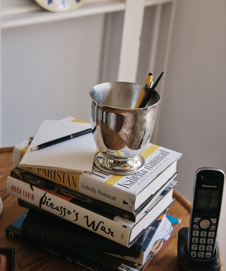 A stack of books, a notepad, a pen, a Lena Pen Pot holding pencils like a pewter vase, and a cordless phone rest on a wooden table.