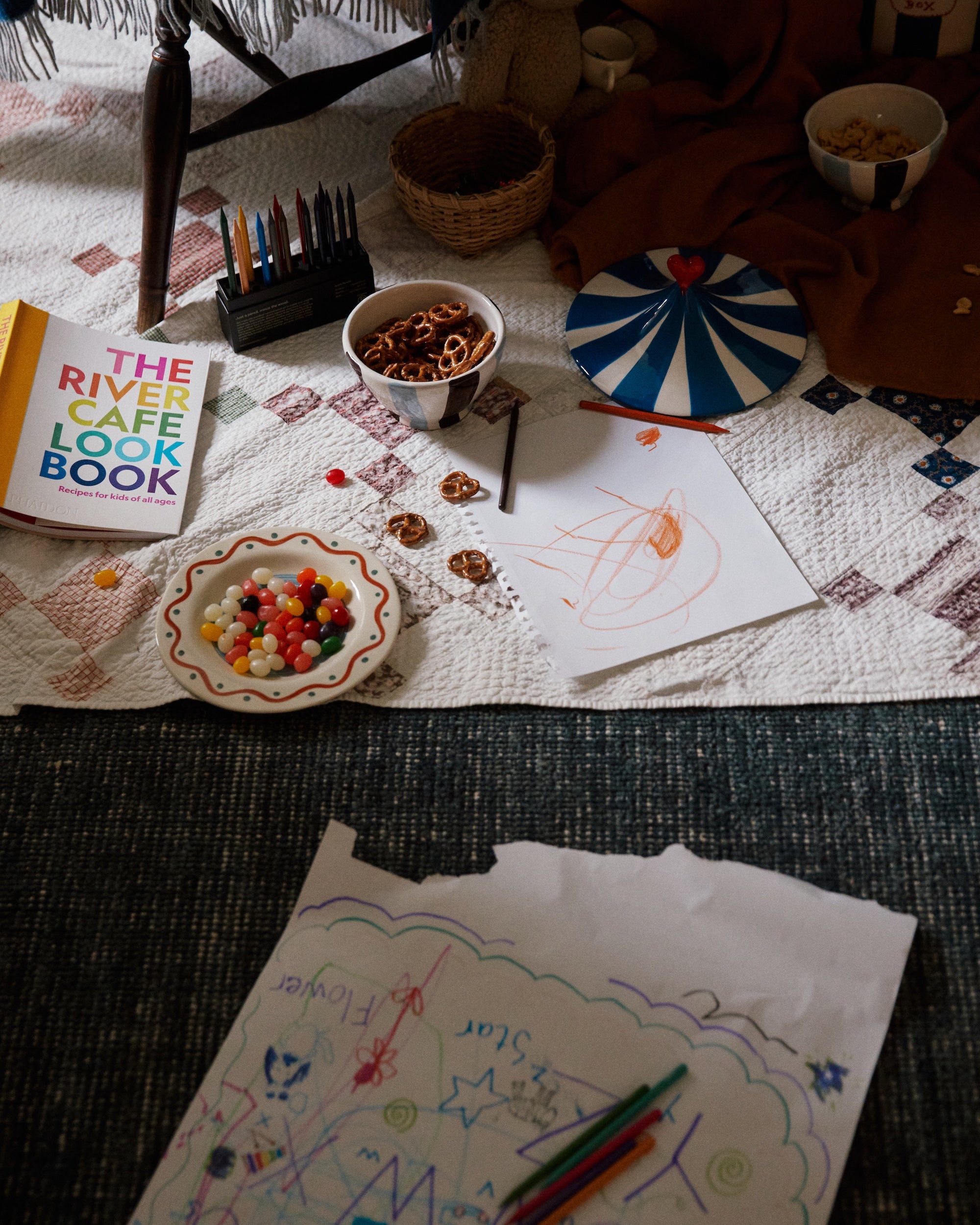 A quilt with snacks, colored pencils, The River Cafe Lookbook: Recipes for Kids, and kids’ drawings spread on the floor next to a table.