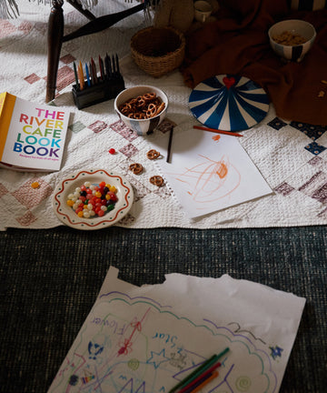 A quilt with snacks, colored pencils, The River Cafe Lookbook: Recipes for Kids, and kids’ drawings spread on the floor next to a table.