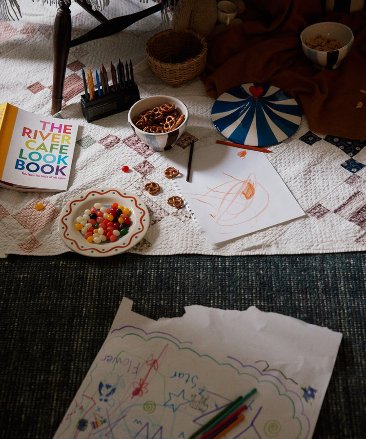 A quilt with snacks, colored pencils, The River Cafe Lookbook: Recipes for Kids, and kids’ drawings spread on the floor next to a table.