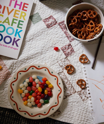 A Colombe Small Plate with jelly beans, a bowl of pretzels, a cookbook, and scribbled paper are arranged on a white quilted surface.