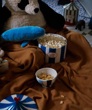 The Love Box sits on a brown blanket beside a circus tent, creating a cozy indoor play or movie setting with plush toys and popcorn.