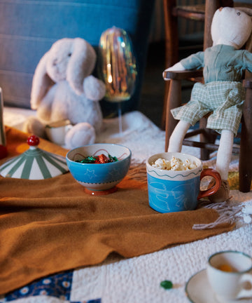 Two whimsical Colombe Mugs, one with popcorn and one with sweets, rest on a blanket beside a stuffed bunny, fabric doll, and festive decorations.
