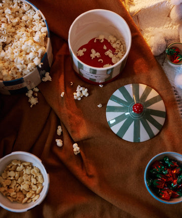 Magic Box tins of popcorn and candies, hand-crafted in Portugal, rest on a brown blanket with scattered popcorn and a striped tin lid nearby.