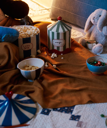 A cozy setup with popcorn bowls, a striped magic container, a stuffed rabbit, and the Ulisse Throw on a patterned surface.