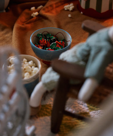 A Colombe Bowl filled with strawberry-shaped candies sits by a cup of popcorn on a blanket near a stuffed doll in a wooden chair.