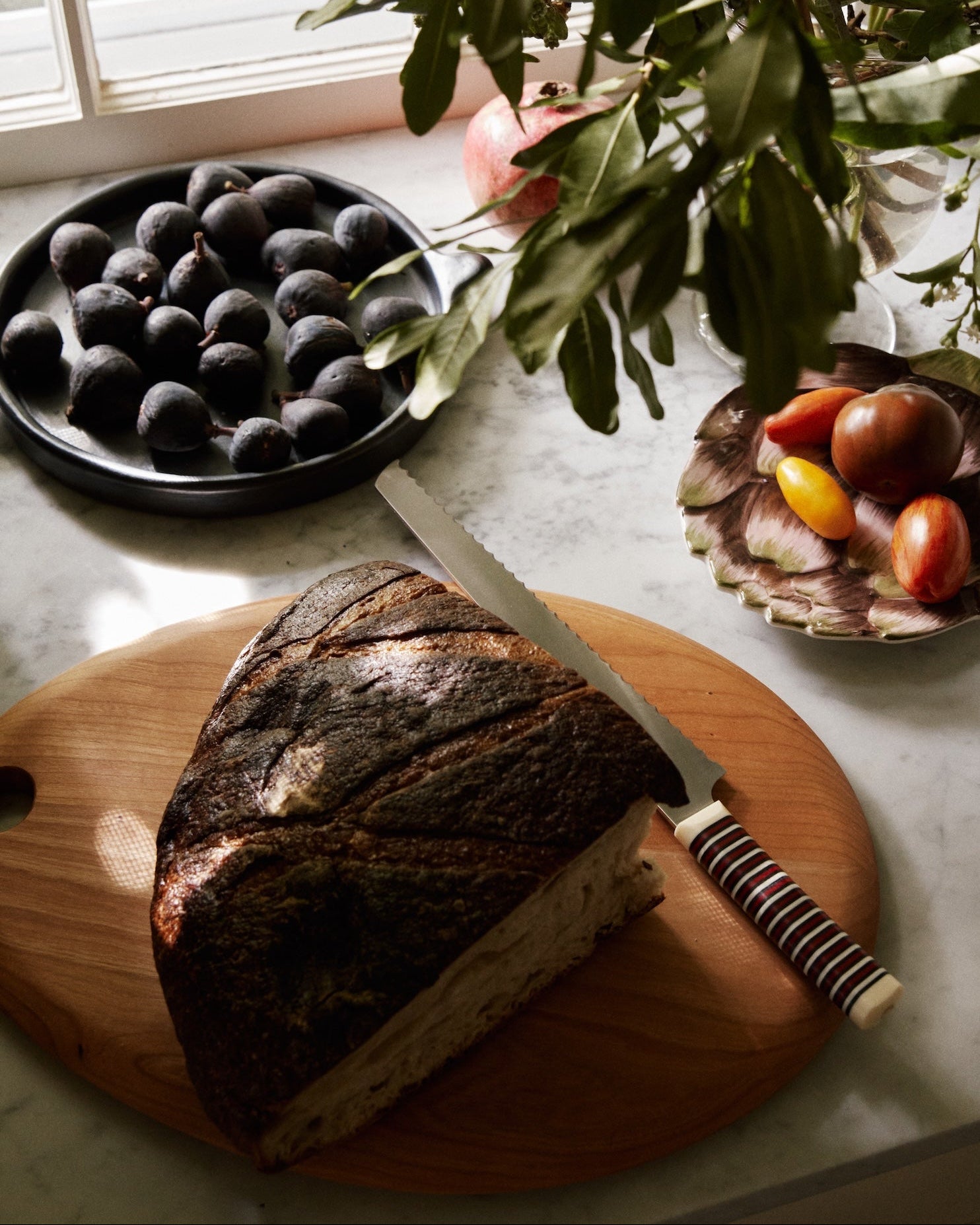 A loaf of bread with a knife rests on the Alden Egg Board, alongside figs and heirloom tomatoes on a marble countertop near a window and leafy plant.