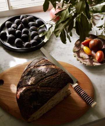 A loaf of bread with a knife rests on the Alden Egg Board, alongside figs and heirloom tomatoes on a marble countertop near a window and leafy plant.