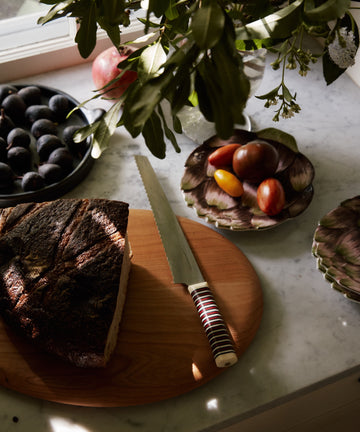 A Bread Knife beside a golden-crusted loaf on a wooden board, with tomatoes and plums on marble under leafy branches.
