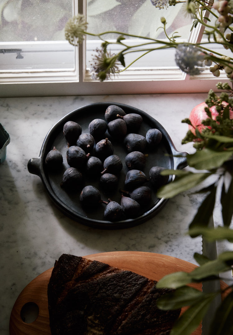 A Chamba Plate filled with fresh figs rests on a marble countertop by a sunlit window, surrounded by flowers and sliced bread on a wooden board.