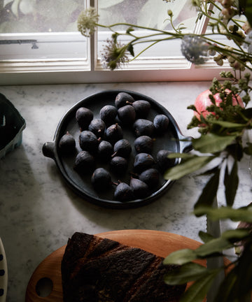 A Chamba Plate filled with fresh figs rests on a marble countertop by a sunlit window, surrounded by flowers and sliced bread on a wooden board.