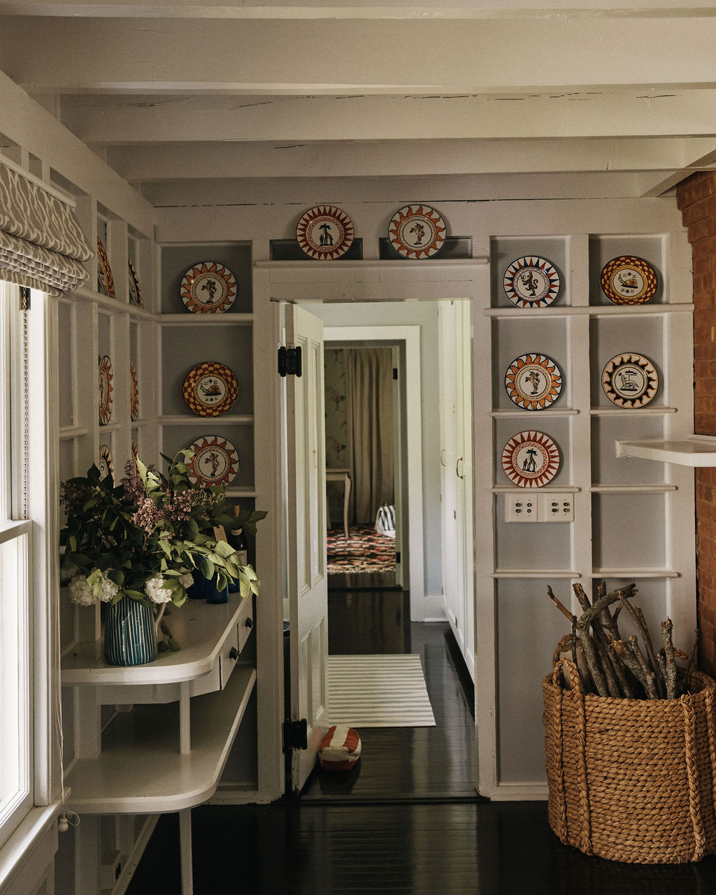 A hallway with white shelves displaying Palio Dinner Plate, The She-Wolf, a table with flower arrangements, a basket of firewood, and an open door leading to another room.