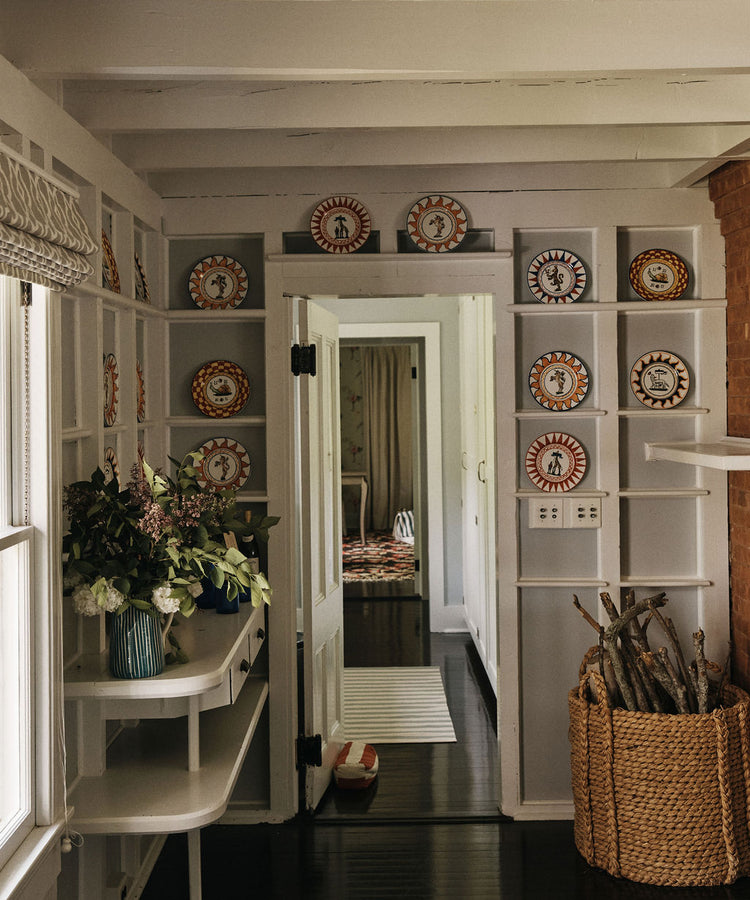 A hallway with white shelves displaying Palio Dinner Plate, The She-Wolf, a table with flower arrangements, a basket of firewood, and an open door leading to another room.