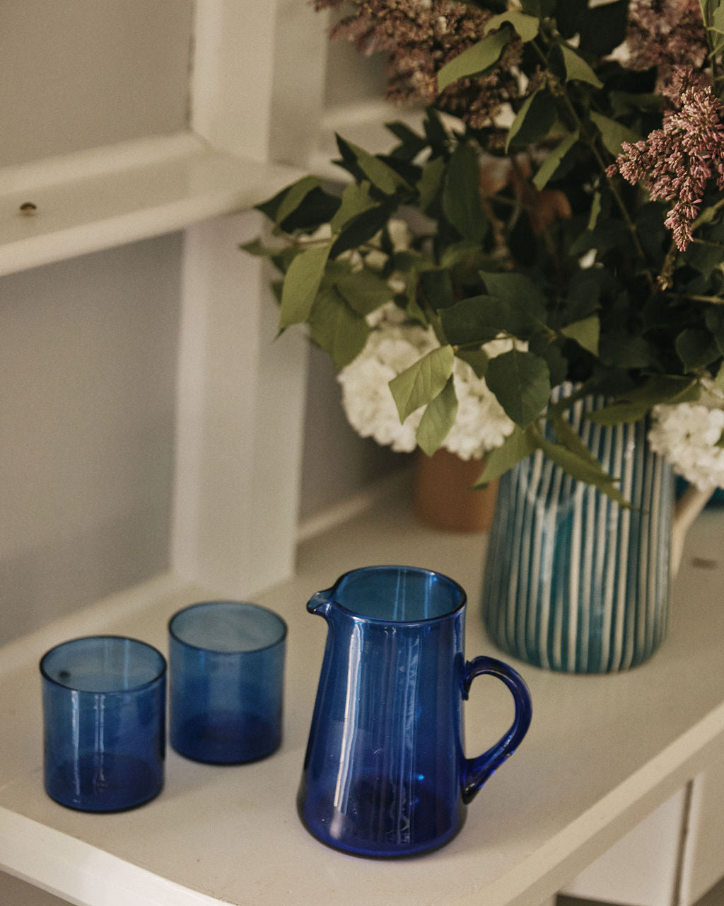 The Elodie Pitcher and two matching blue glass cups sit on a white shelf beside a flower-filled vase, bringing a hint of Damascus charm.