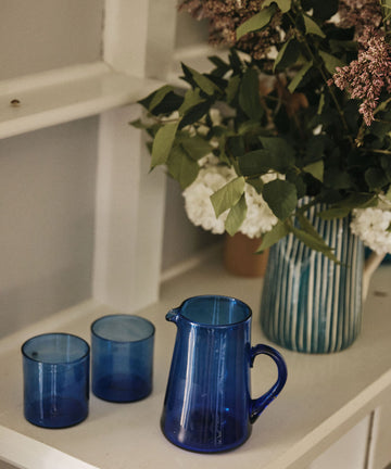 The Elodie Pitcher and two matching blue glass cups sit on a white shelf beside a flower-filled vase, bringing a hint of Damascus charm.