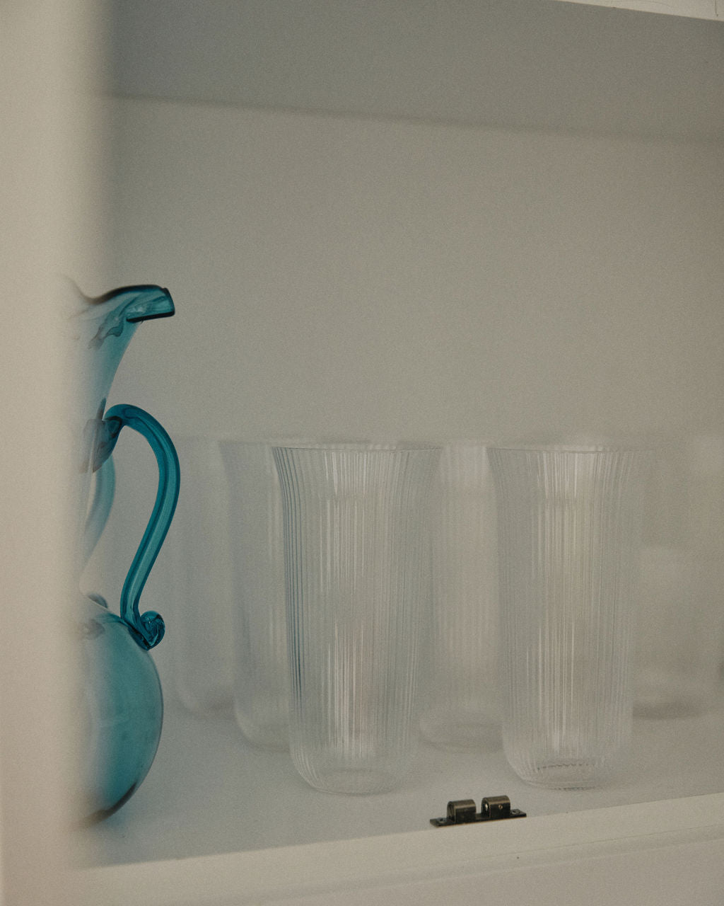 A blue glass pitcher sits beside the Linea Ribbed Water Glass, Set of 4, and sturdy glassware, evoking a coastal bistro vibe inside the sleek white cabinet.