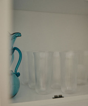 A blue glass pitcher sits beside the Linea Ribbed Water Glass, Set of 4, and sturdy glassware, evoking a coastal bistro vibe inside the sleek white cabinet.