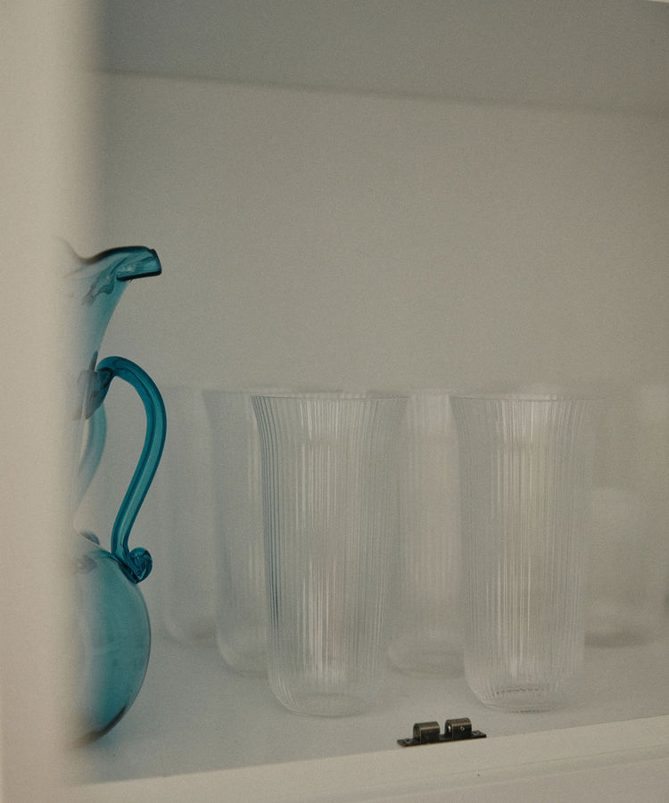A blue glass pitcher sits beside the Linea Ribbed Water Glass, Set of 4, and sturdy glassware, evoking a coastal bistro vibe inside the sleek white cabinet.