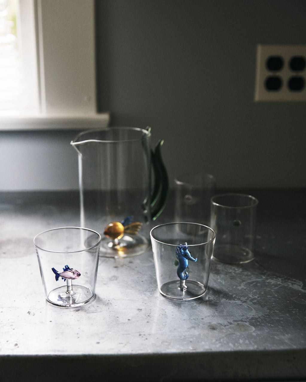 The Lido Tumbler, Set of 4—borosilicate glasses with small animal figurines—sits on a grey countertop near a window and wall outlet.
