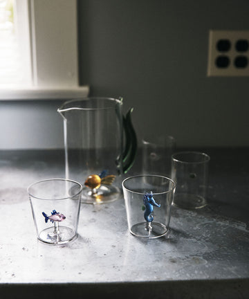 The Lido Tumbler, Set of 4—borosilicate glasses with small animal figurines—sits on a grey countertop near a window and wall outlet.