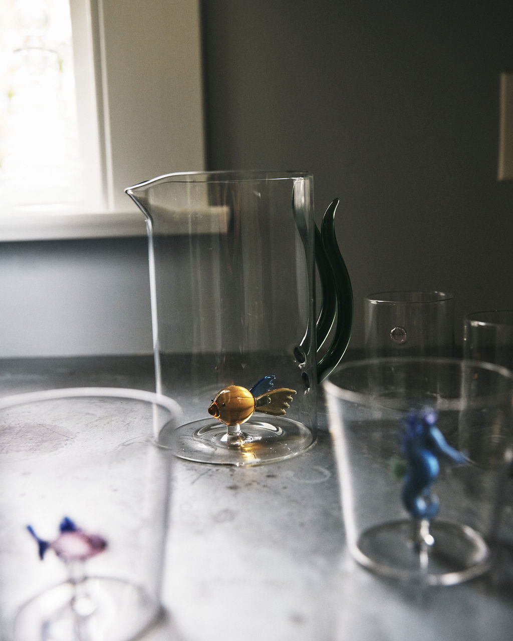 The Fish Pitcher, a unique hand-blown glass piece with a green handle and a small glass fish at its base, sits on the table. In the foreground are two clear glasses, each featuring a colorful glass fish inside.