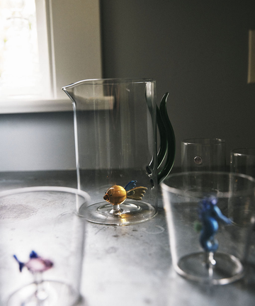 The Fish Pitcher, a unique hand-blown glass piece with a green handle and a small glass fish at its base, sits on the table. In the foreground are two clear glasses, each featuring a colorful glass fish inside.