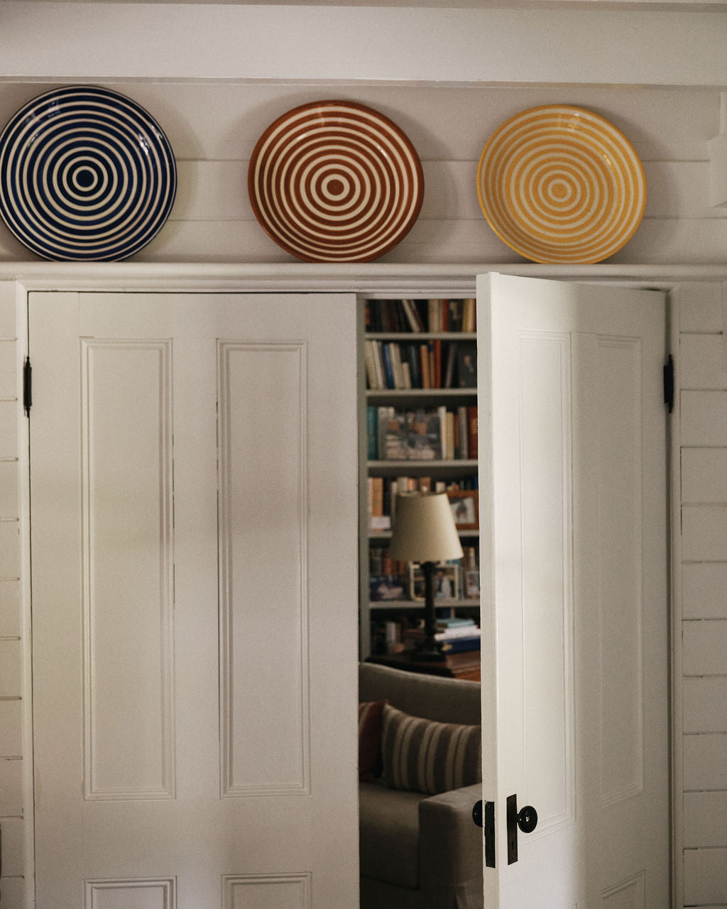 Three Natalia Platter decorative plates, each featuring unique concentric circle patterns, are displayed above white double doors that are slightly open to reveal a room with bookshelves and a lamp.