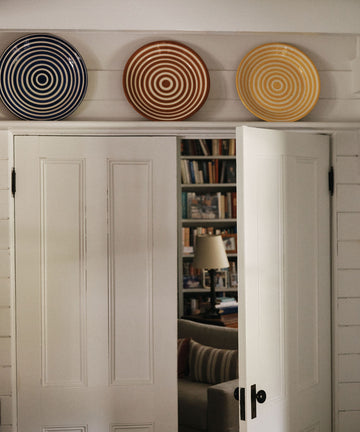Three Natalia Platter decorative plates, each featuring unique concentric circle patterns, are displayed above white double doors that are slightly open to reveal a room with bookshelves and a lamp.