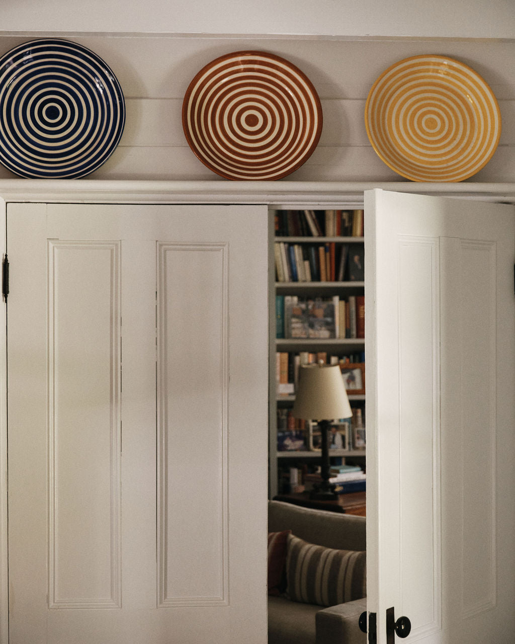 The Natalia Platter, with its blue, brown, and yellow concentric circles, hangs alongside two other decorative plates above double doors, partially open to show a room with bookshelves and a lamp.
