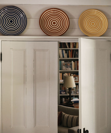 The Natalia Platter, with its blue, brown, and yellow concentric circles, hangs alongside two other decorative plates above double doors, partially open to show a room with bookshelves and a lamp.