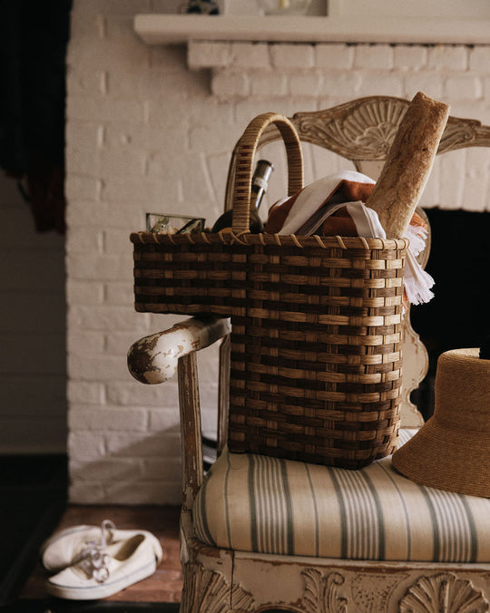 A handwoven Stair Tote filled with a loaf of bread, a glass jar, and a cloth rests on a striped chair beside a straw hat, while white shoes are placed on the floor nearby.