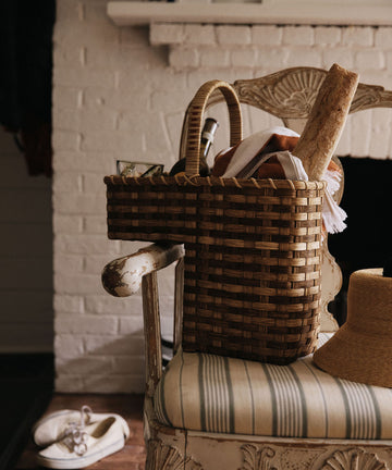 A handwoven Stair Tote filled with a loaf of bread, a glass jar, and a cloth rests on a striped chair beside a straw hat, while white shoes are placed on the floor nearby.