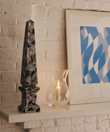 A blue and white obelisk, the Lauren Hurricane Lamp with a lit candle, and a framed blue abstract print rest on a white shelf against a white brick wall.