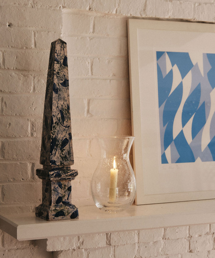 A blue and white obelisk, the Lauren Hurricane Lamp with a lit candle, and a framed blue abstract print rest on a white shelf against a white brick wall.