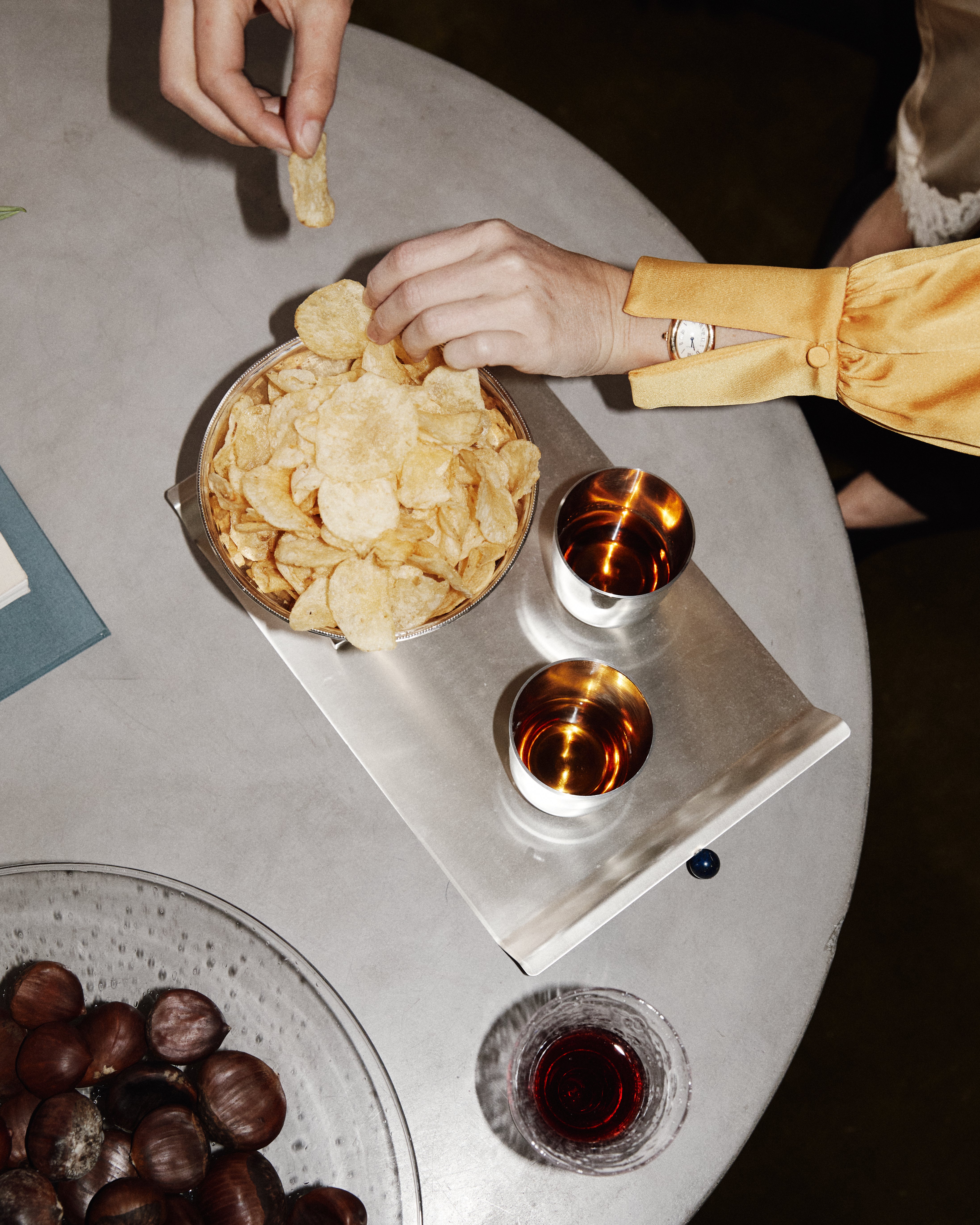Two hands reach for potato chips from a bowl on the Roberta Serving Tray, set atop a round table with two drinks, chestnuts, and a glass of red beverage.