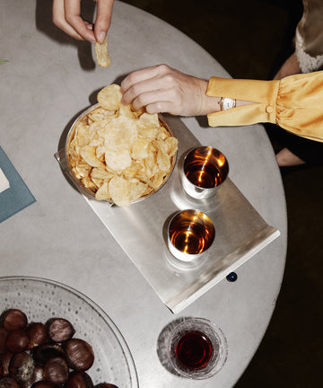 Two hands reach for potato chips from a bowl on the Roberta Serving Tray, set atop a round table with two drinks, chestnuts, and a glass of red beverage.