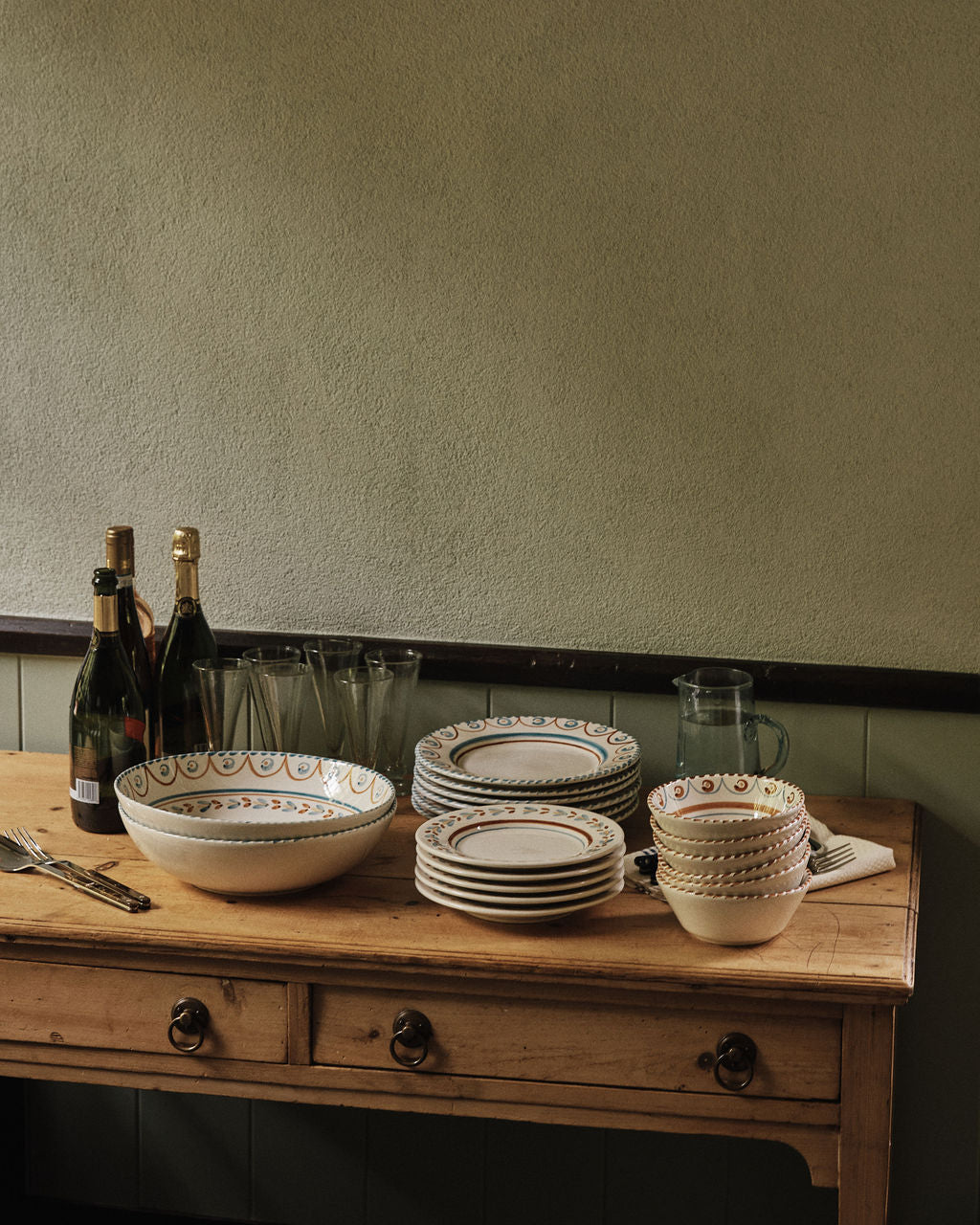 A wooden table holds stacked bowls and plates, including an Ottoline Serving Bowl, drinking glasses, a pitcher, cutlery, and three bottles of sparkling wine against a plain wall.